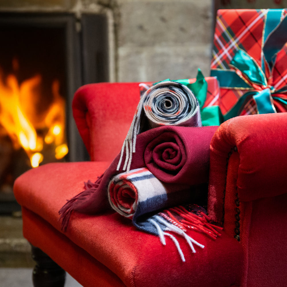 Selection of rolled cashmere scarves in plaid and plain designs on a red armchair in front of a roaring fireside