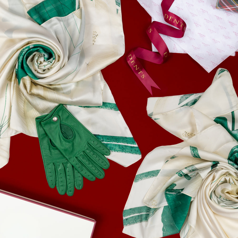 Women's green leather driving gloves and two silk-like women's scarves with white and green patterns set against a red background with Dents gift ribbon and gift box