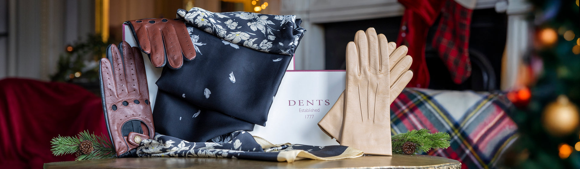 Men's brown leather driving gloves and women's beige leather gloves with a women's silk-like floral scarf and gift box on a table with a festive background