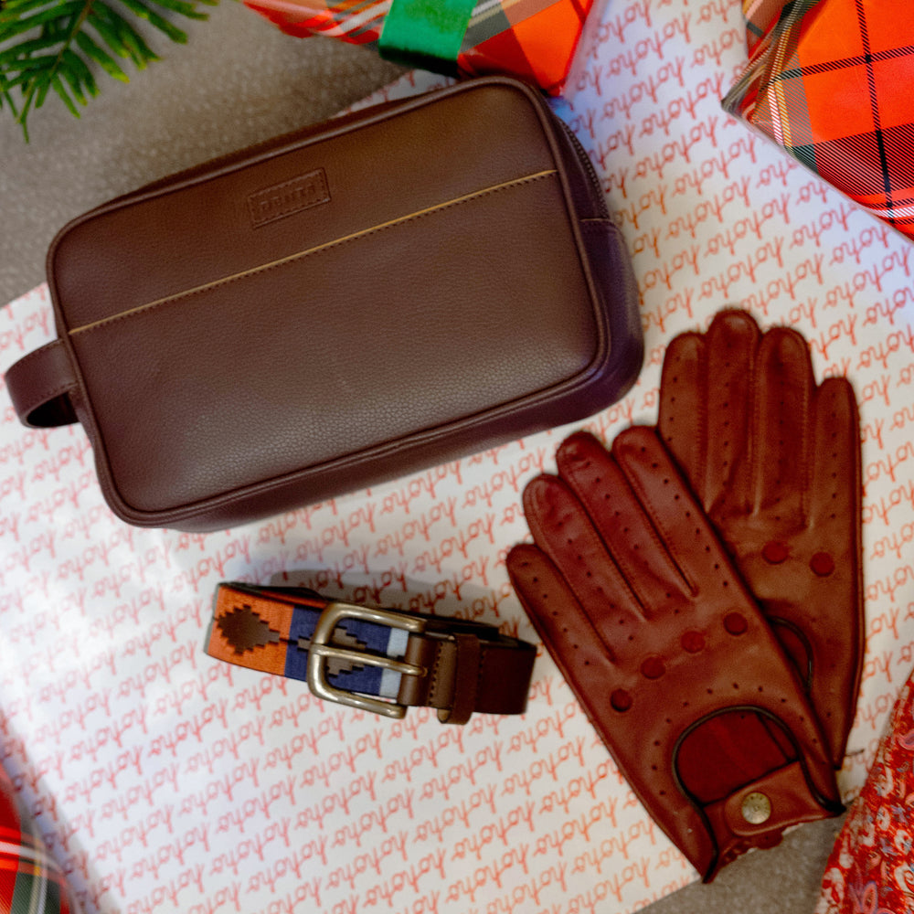 A men's brown leather washbag, pair of brown leather driving gloves and a blue and brown polo belt on top of Christmas wrapping paper and with wrapped gift boxes