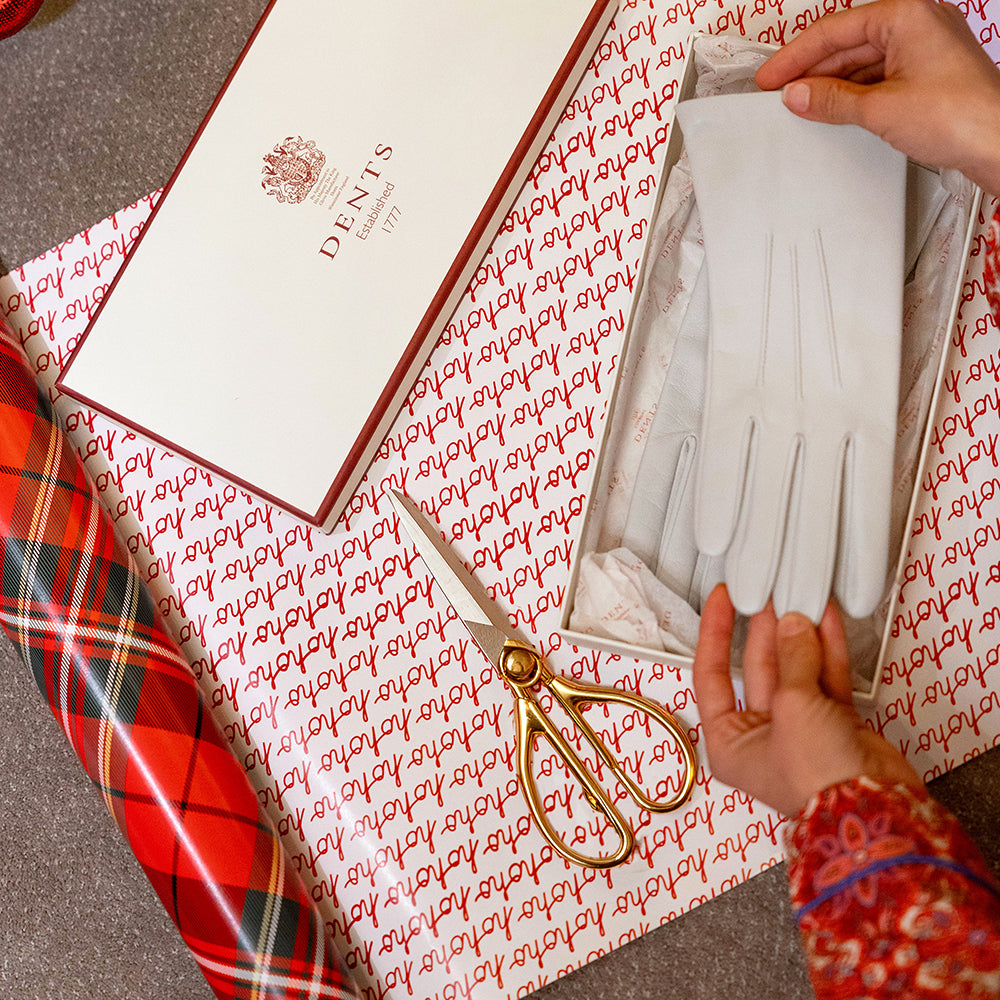 Close-up on women's hands placing a pair of women's white leather gloves into a Dents gift box, with gold scissors and wrapping paper