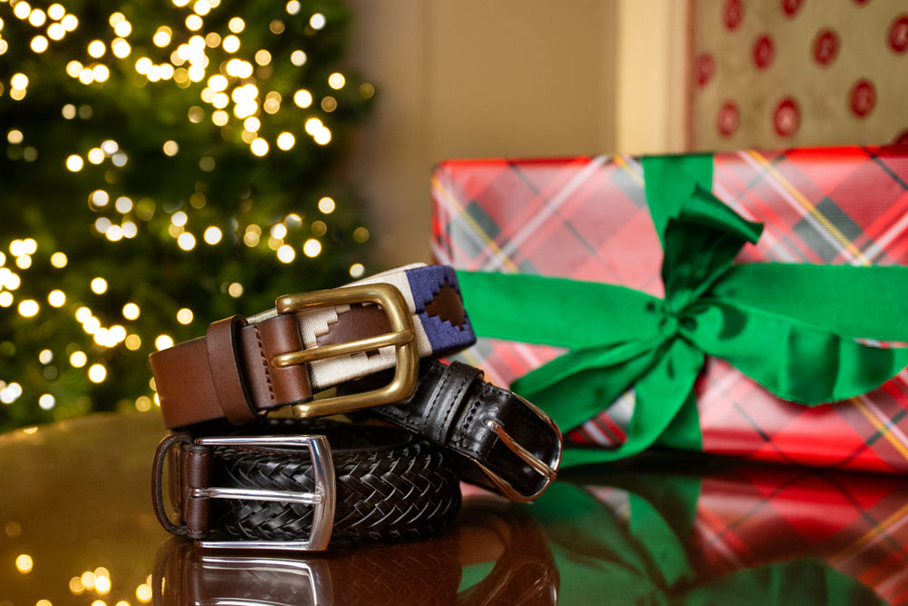 Three men's leather belts stacked on a table next to a wrapped Christmas present and with a Christmas tree in the background