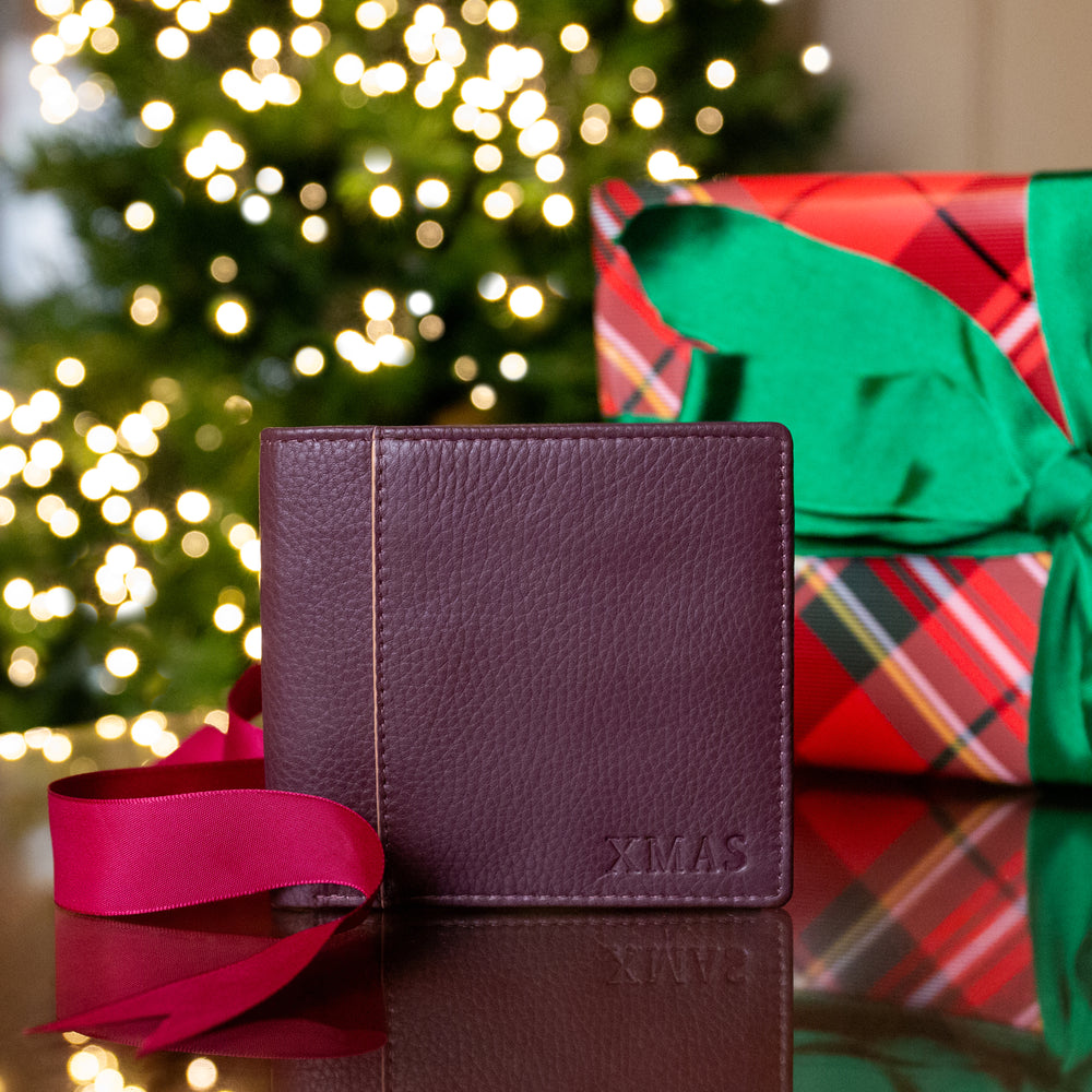 Men's brown leather bifold wallet embossed with 'XMAS' on a table with a ribbon and wrapped Christmas presents with a Christmas tree in the background