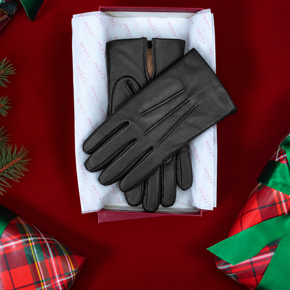 Pair of men's black touchscreen leather gloves on top of tissue paper in a Dents gift box against a red background with wrapped presents