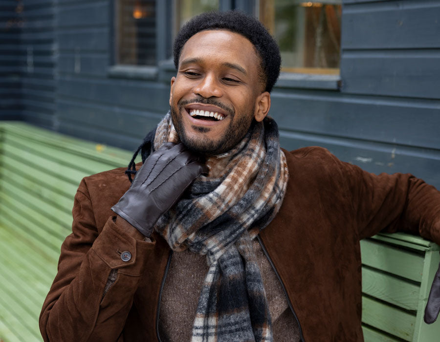 Man wearing blanket scarf and three-point leather gloves in brown, sitting outside.