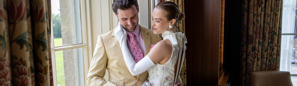 Woman wearing long satin gloves in white in a wedding dress standing with a man by a window