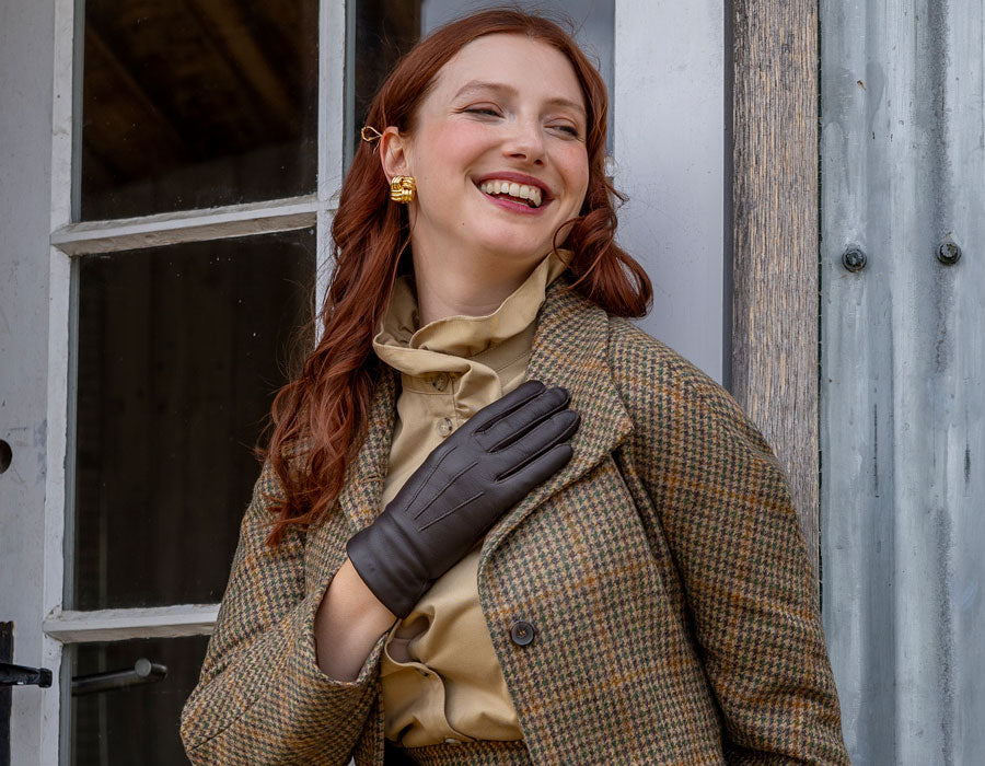 Woman wearing three-point leather gloves in brown, leaning against a door.
