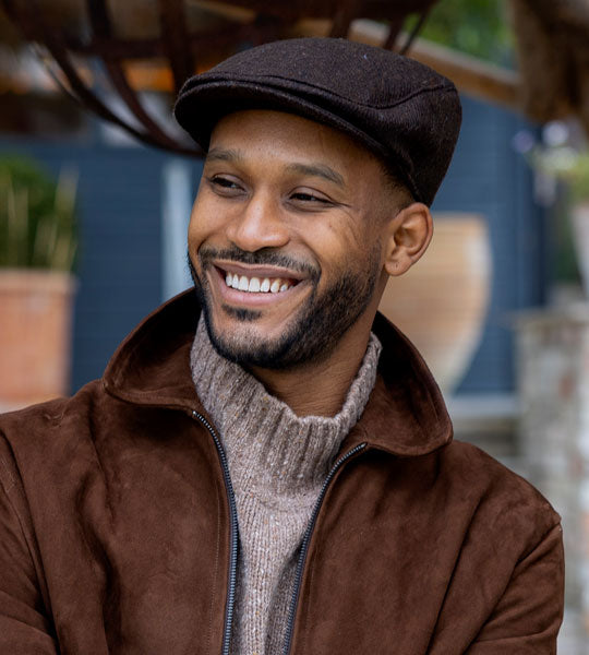 man wearing brown flat cap with brown coat outside next to trees.