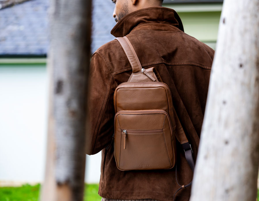 Man wearing small brown leather backpack with brown jacket in trees.