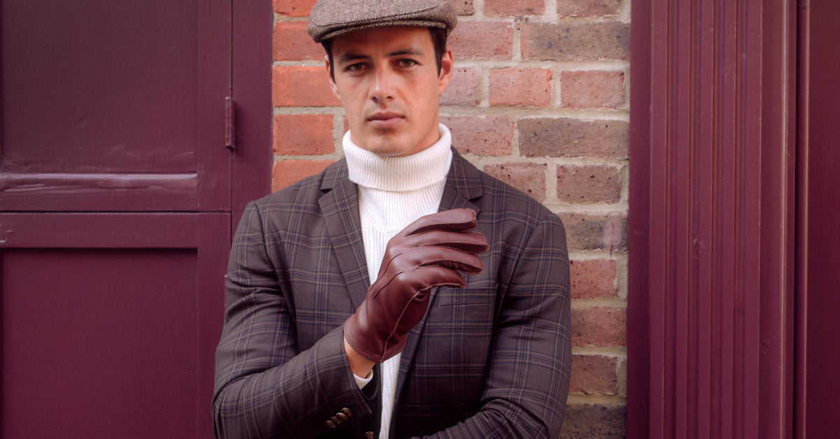 Man wearing three-point wool-lined leather gloves in tan, leaning against a wall. 