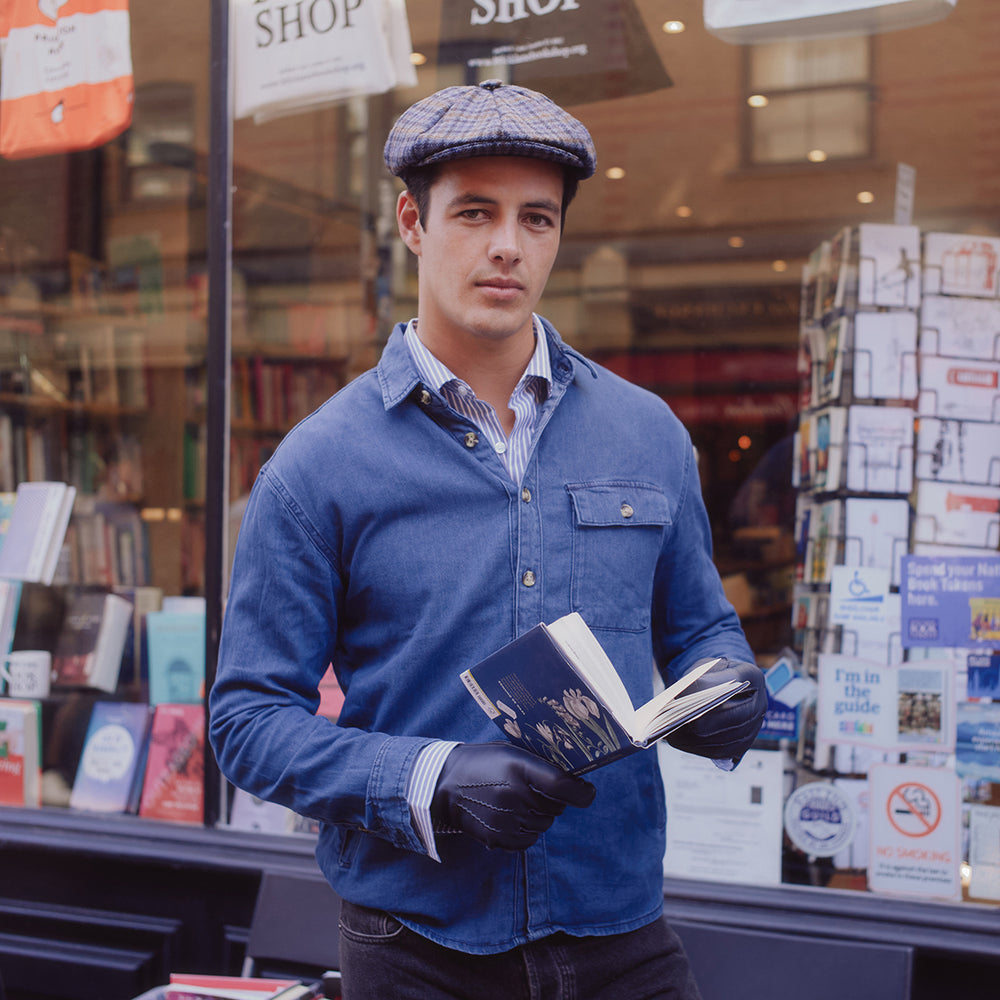 Man wearing a tweed hat and blue leather gloves reading a book outside a bookshop