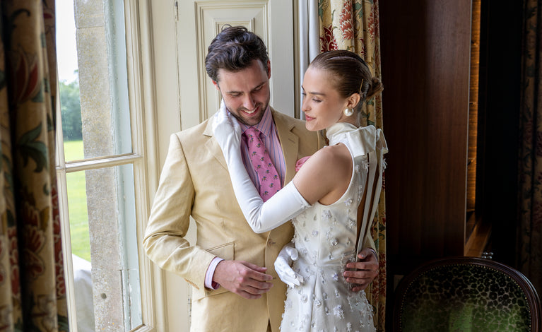 Woman wearing long satin gloves in white in a wedding dress standing with a man by a window