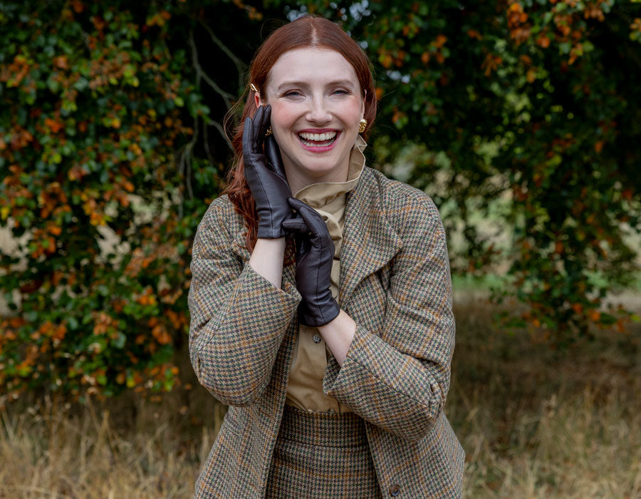 woman wearing brown three-point leather gloves with tweed jacket in a field. 