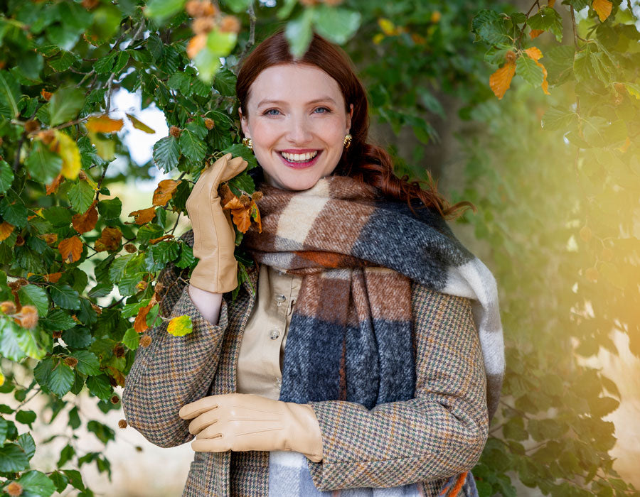 Woman wearing checked blanket scarf in oranges and beige with beige leather gloves, underneath a tree.