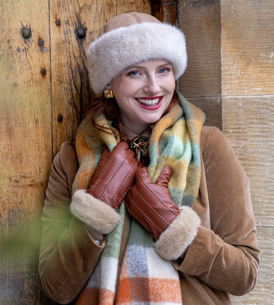 Woman wearing blanket scarf, leather gloves with faux fur cuffs and a faux suede hat, leaning against a door. 