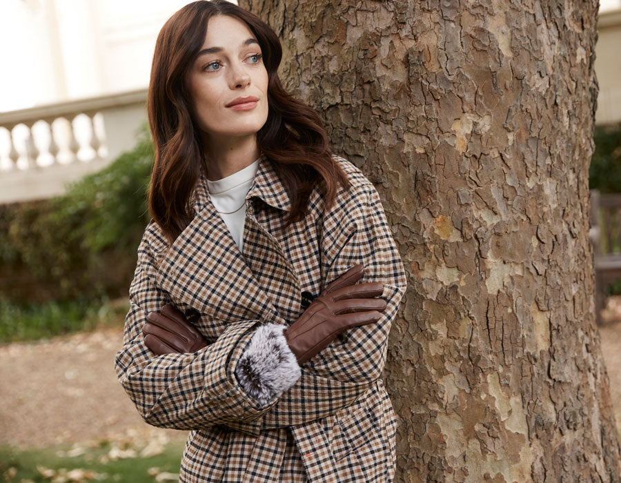 A woman wearing chestnut leather gloves with faux fur cuffs by a tree