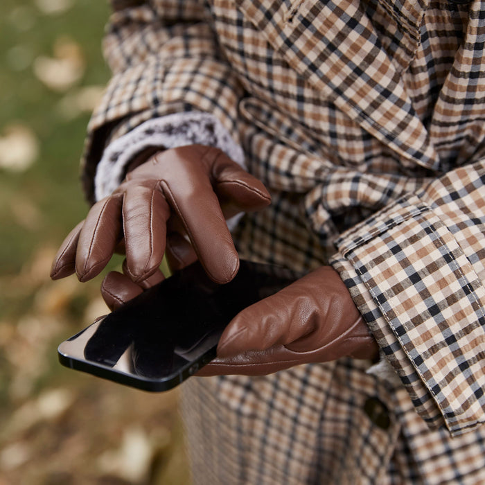 Woman wearing leather touchscreen gloves with faux fur cuffs in chestnut using her phone at the park